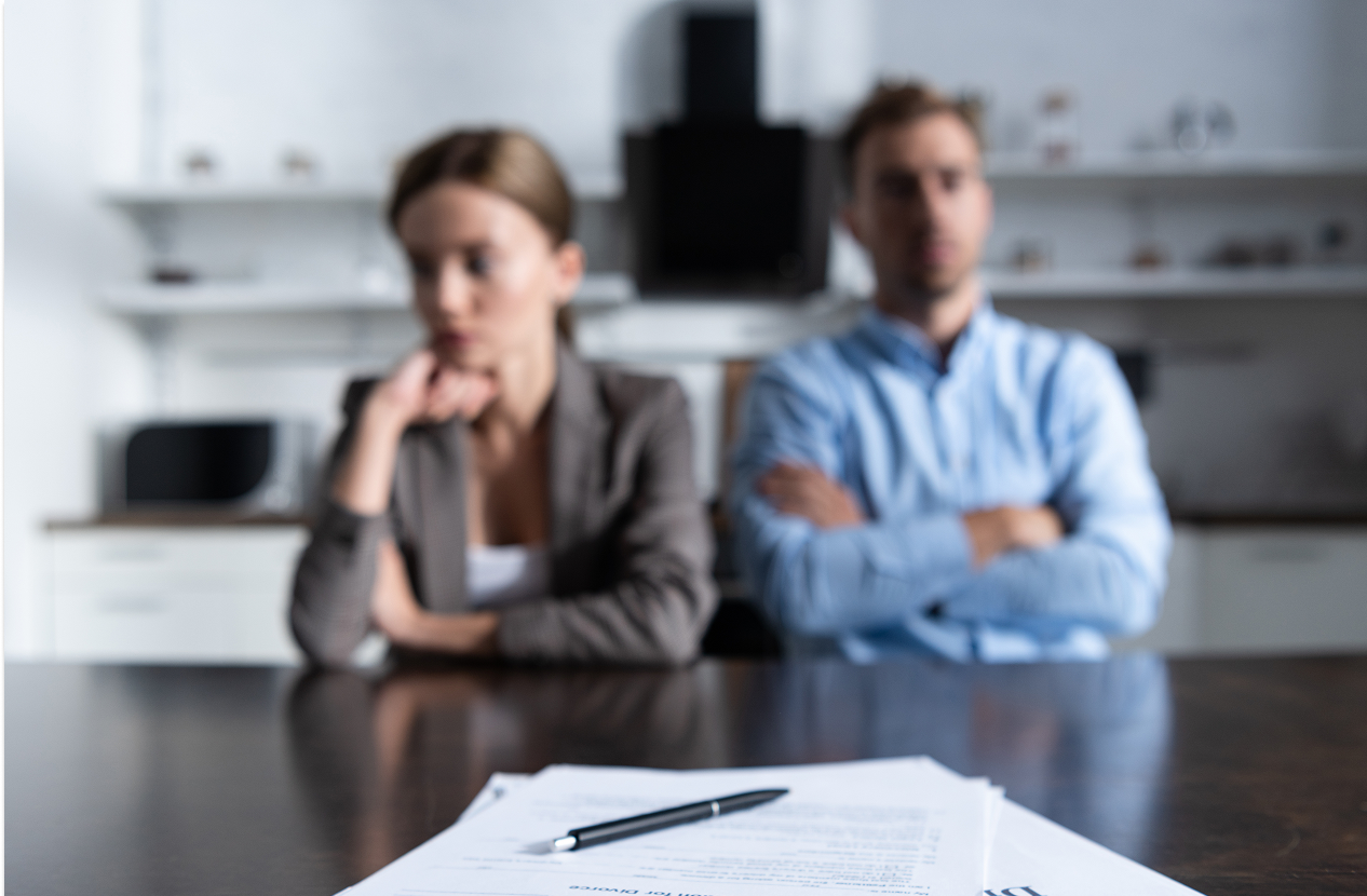 Pen on divorce petition documents; blurred woman looking down and man with crossed arms.