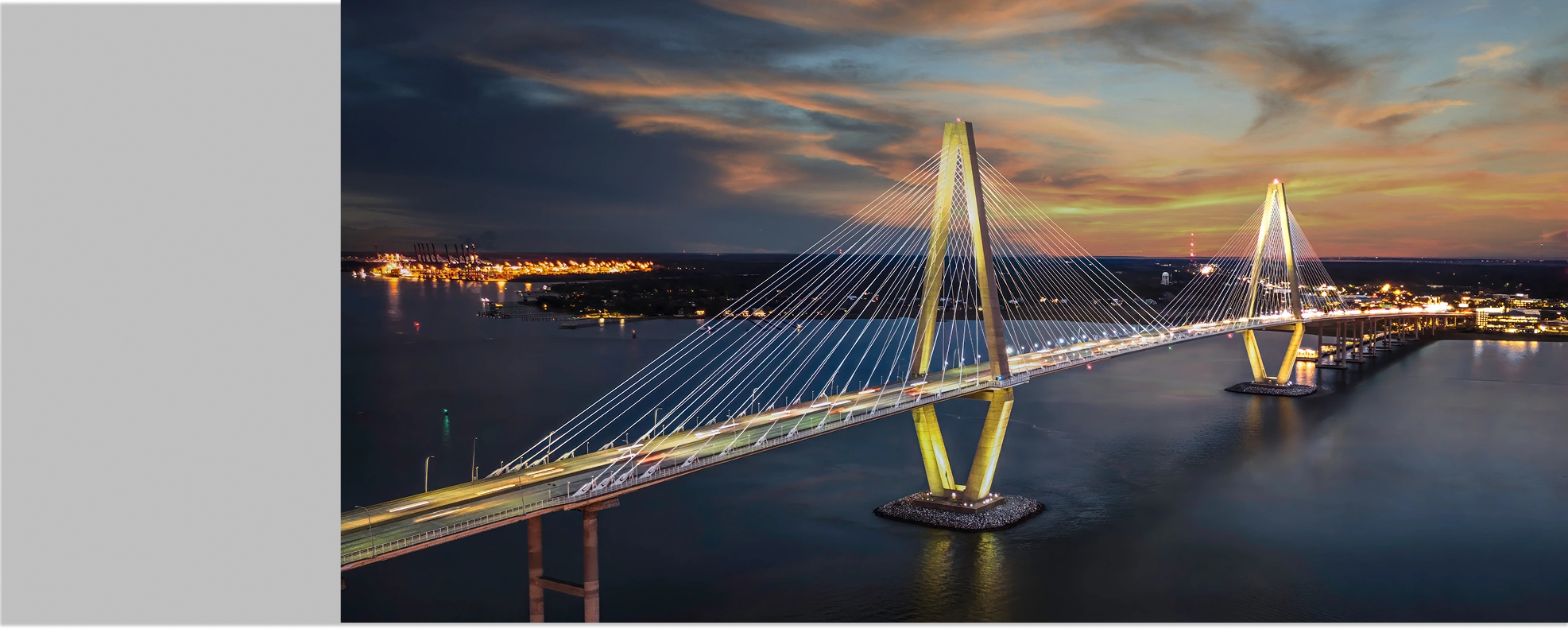 Illuminated cable-stayed bridge over water at sunset with light trails from cars and city lights in the distance.