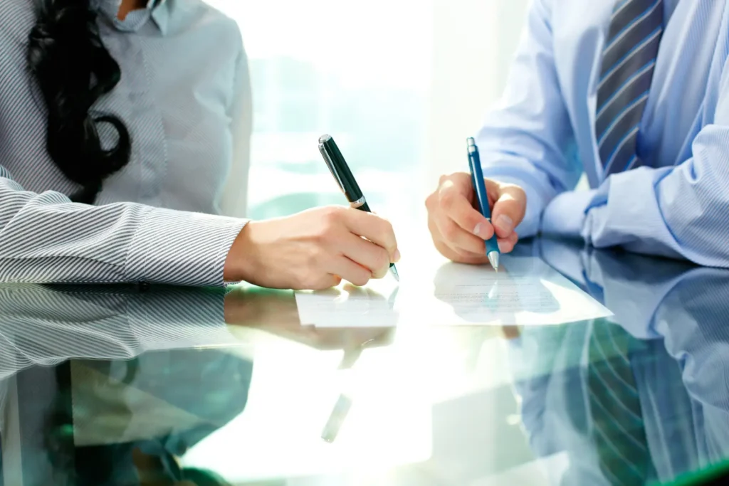 Two people's hands sign documents with pens on a reflective glass table, one in a striped shirt, the other in a blue shirt.