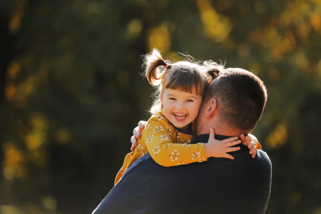 Smiling young girl with pigtails hugs a parent outdoors, bathed in warm sunlight.