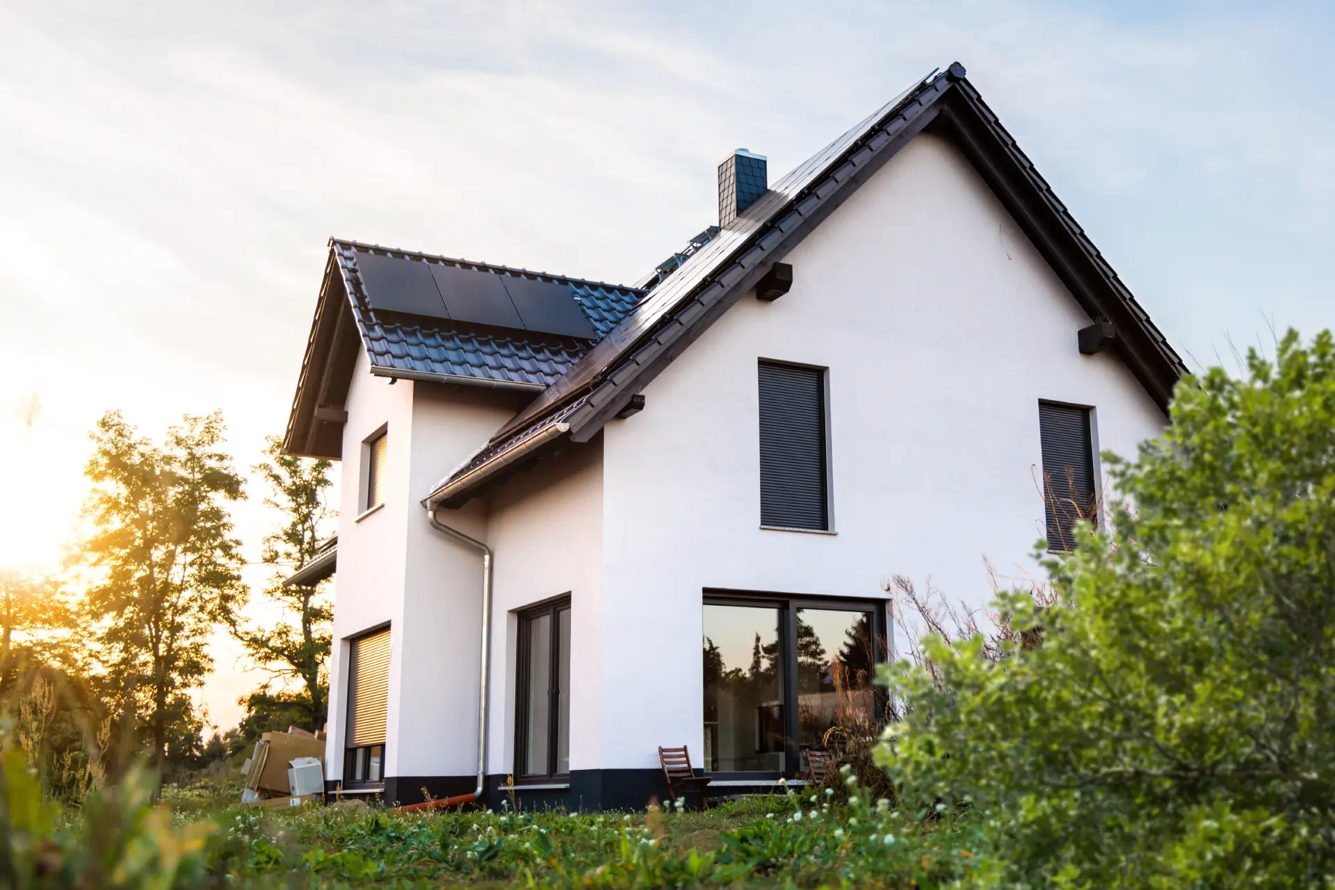 Modern white house with a dark tiled roof, solar panels, and window shutters, bathed in golden sunlight.